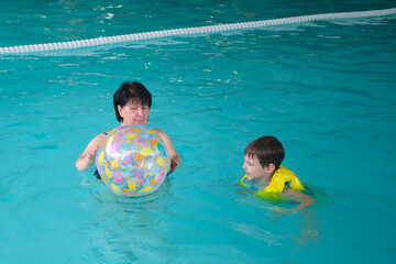 Woman coaching a boy to swim in an indoor pool with a colorful beach ball. Learning to swim and water safety concept. A child aged four years.