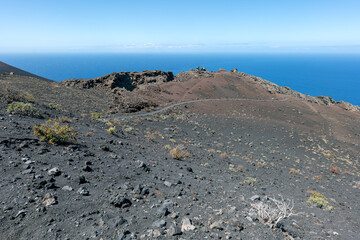 Volcanic landscape with black lava rock, sparse vegetation and a hiking trail – captured in southern La Palma with ocean view under a clear sky.