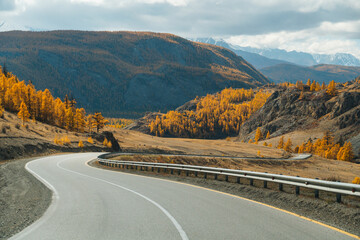A view of a winding road with a guardrail leading into a scenic mountain valley with hillsides covered in yellow autumn trees