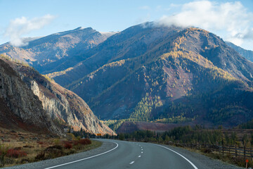 A winding road leads into a valley with large mountains covered in autumn trees and a mix of sunshine and clouds in the sky