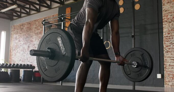 African American man in twenties in gym spotting loaded barbell, gripping and performing deadlift