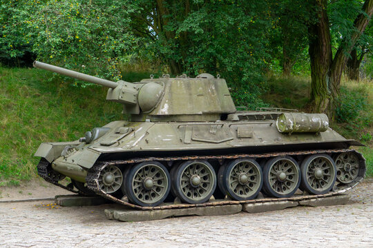 Soviet T-34 medium tank parked on a cobblestone surface, surrounded by trees, side view with clearly visible tracks and turret