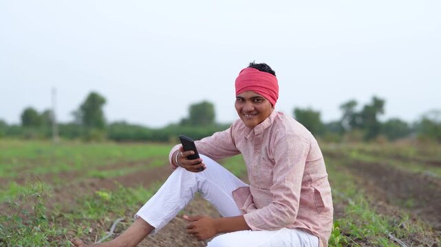 young indian farmer holding smartphone sitting at green agricultural field	