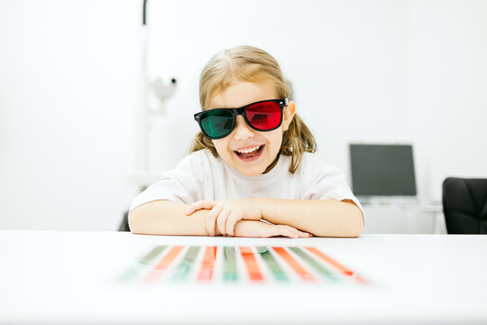 Young girl wearing colorful vision testing glasses, smiling joyfully while sitting at a table with eye chart, showcasing a playful atmosphere in an ophthalmology clinic setting