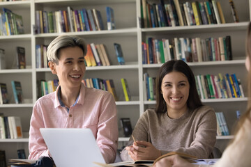 Friendly scholar community. Smiling diverse teen pair of students study together with groupmates in library engaged in learning discussion using books digital tools laugh chat have fun enjoy good joke