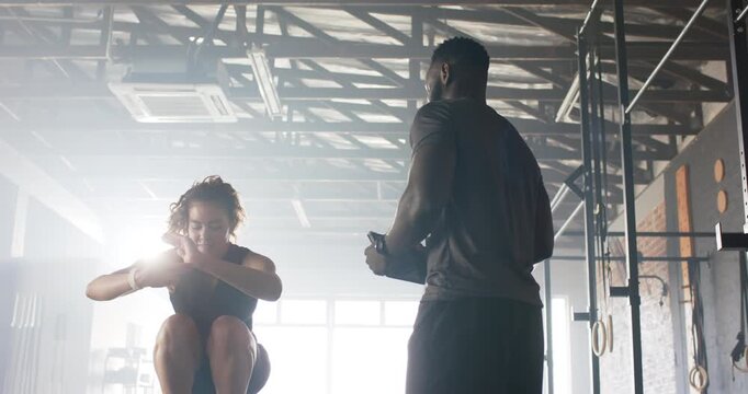 African American trainer tapping stopwatch and female athlete performing squat jumps at gym