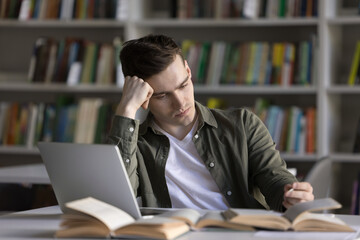 Academic challenge. Tired serious young man university student learning remotely at library desk using printed books and online information sources think hard on difficult problem solution holding pen