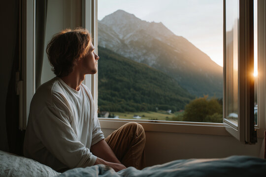 Thoughtful young man sitting by an open window, gazing at the serene mountain landscape during sunset