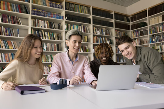 Collaborative remote studying. Group of 4 smiling motivated multicultural young people college university students meet at library desk to watch video lecture on online education platform via notebook