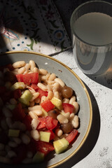 Ensalada de alubias blancas con tomate, pepino y cebollitas encurtidas en vinagre de Módena con un vaso de limonada, sobre plato gris y fondo blanco