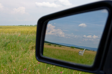 A scenic car mirror reflection of a bench on a grassy field under a beautiful, bright sky.