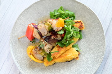 Top-down view of a colorful plated meal featuring a grilled chicken skewer, fresh salad, and creamy polenta on a white background.