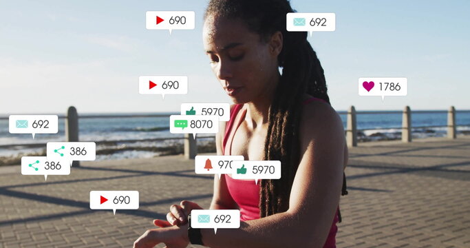 Standing on seafront walkway, athletic woman in red top checking smartwatch and social media icons