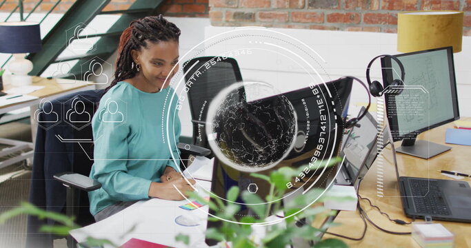 Woman in teal blouse sketching chart and viewing code on laptop in open-plan office, with monitors