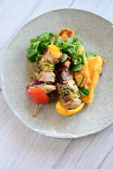 Top-down view of a colorful plated meal featuring a grilled chicken skewer, fresh salad, and creamy polenta on a white background.