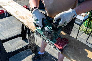 A woman grinds a board with a belt sander on trestles in the garden.