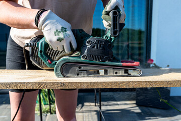 A woman grinds a board with a belt sander on trestles in the garden.