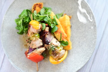 Top-down view of a colorful plated meal featuring a grilled chicken skewer, fresh salad, and creamy polenta on a white background.