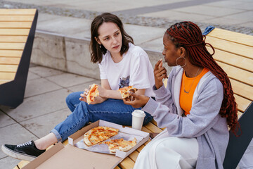 Two young women having pizza and talking on city bench
