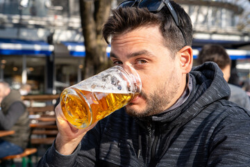 Hombre con gafas tomando una cerveza helles mientras mira a cámara, en un patio cervecero en Munich, Alemania