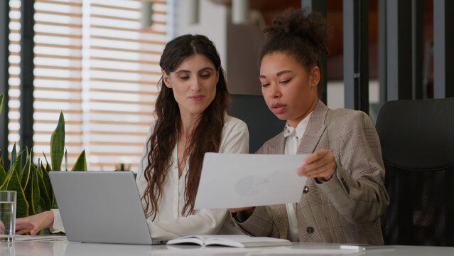 African American Caucasian businesswomen two female coworkers multiethnic business partners colleagues girls women together discuss papers graphs sales data use laptop teamwork in office partnership