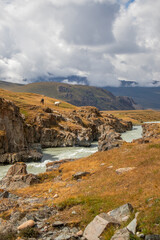 Obraz premium Two horses, one black and one white, stand on a grassy hill in a rocky mountain gorge with a river flowing below and a cloudy sky