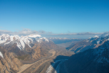 A panoramic aerial view of a vast mountain valley with a large glacier, surrounded by rugged snowcapped peaks and a clear blue sky