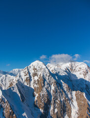 A vertical shot of rugged, rocky, snowcapped mountains under a clear blue sky with some small clouds in the distance