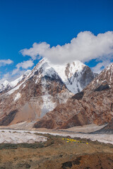 A vertical view of a high-altitude mountaineering camp with yellow tents in a rocky landscape with a snowcapped mountain peak in the background