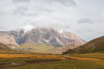 Panoramic view of majestic snowcapped and rocky mountain peaks towering over a vast, golden and green valley under a dramatic cloudy sky