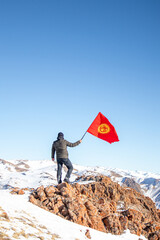 Obraz premium A man in winter clothing stands on a rocky mountain peak, holding a waving red flag of Kyrgyzstan with a vast snow-covered mountain range in the background