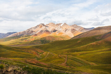 A wide view of a vast, golden and green mountain valley with a serpentine dirt road, leading towards majestic peaks under a cloudy sky