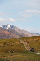 Fototapeta premium An off-road vehicle drives on a dirt road through a grassy valley, with majestic snowcapped mountains in the background under a blue sky