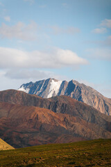A panoramic view of a rugged, multi-colored mountain range with snowcapped peaks under a clear blue sky with some clouds