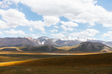 A panoramic view of a vast, golden and green valley with majestic snowcapped mountains in the background under a bright blue sky with clouds