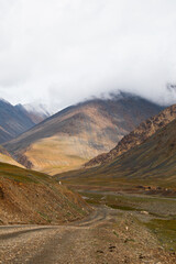 A vertical shot of a winding gravel road leading through a mountain valley with green and brown hills, partially covered by low clouds