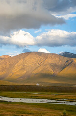 A traditional white yurt sits in a vast golden and green valley with a large, sunlit mountain in the background under a beautiful cloudy sky