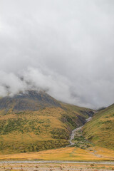 A vertical shot of a river flowing from a mountain peak shrouded in low clouds, down a grassy slope into a golden valley