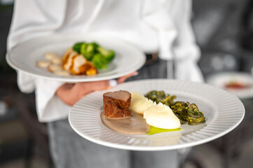Close-up of two white plates with assorted cooked meals including mashed potatoes, meat, broccoli and greens, presented in a well-lit dining setting.