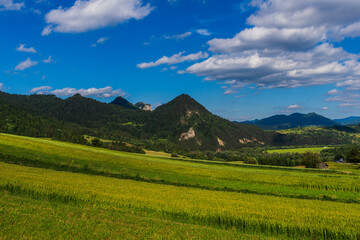View of layered green fields and tree-covered hills with sharp rocky peaks in the background and clouds floating over the clear blue sky.
