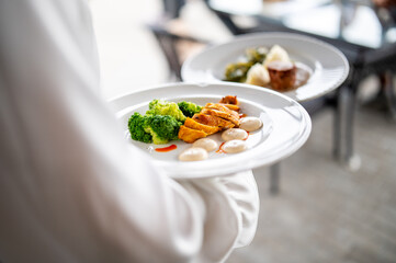 Waiter serving a gourmet meal with sliced chicken, broccoli, and sauce on a white plate, presented in a restaurant setting with elegant food styling.