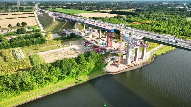 Aerial footage of highway bridge construction over waterway. Highway bridge project under construction including construction crane, support pillars and traffic on the existing bridge.