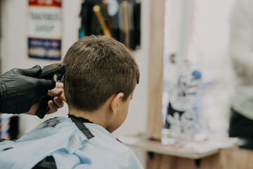 Fototapeta premium Smiling little boy sitting at barber shop, while hairdresser styling his hair.