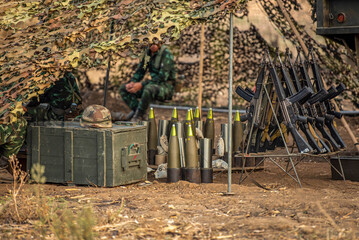 Crew and Ordnance Under Camouflage, Military personnel under camouflage netting beside a rack of rifles and artillery shells, preparing munitions and weapons in a forest field training advance.