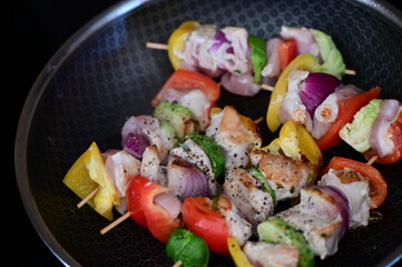 Raw chicken skewers with colorful vegetables on a black frying pan, set against the background of meal preparation