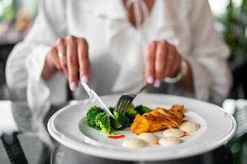 Person cutting cooked broccoli and grilled chicken with sauce on plate, close-up view of healthy meal on table