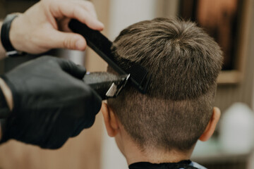 Fototapeta premium Smiling little boy sitting at barber shop, while hairdresser styling his hair.