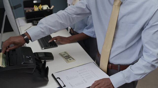 Tilt down shot of young black male employee inserting paper cash into automated counting machine while female colleague working on computer in bank office