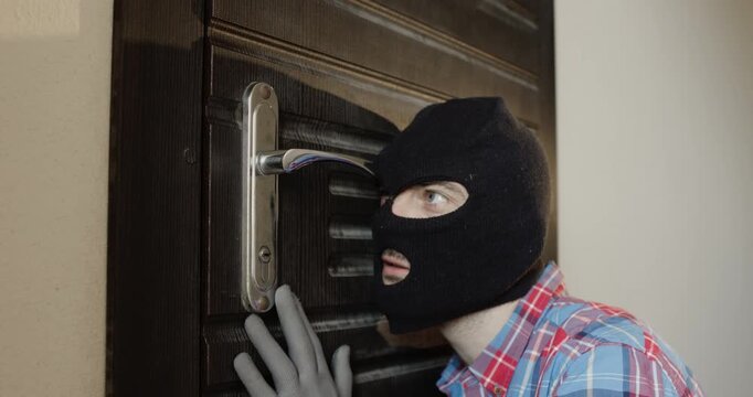 Man in black balaclava and gloves pulls doorknob with quick motion. Dangerous burglar presses ear against door of apartment and tries to hear footsteps or voices