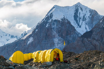 A line of bright yellow tents on a rocky ridge with a breathtaking view of a majestic snowcapped mountain peak under a clear blue sky